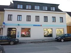 Ein weißes Gebäude mit einem blauen Streifen oben und Fenstern im ersten Stock. Zwei schwarze Autos sind vor dem Gebäude geparkt.