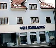 Ein weißes Gebäude mit roten Dachziegeln und mehreren Fenstern. Das Gebäude hat ein Schild, das 'Volksbank' in fetten Buchstaben liest.