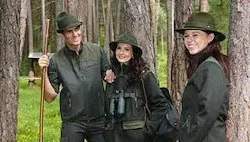 Drei Personen, die in grüne Kleidung und Hüte gekleidet sind, stehen in einem Wald. Der Mann auf der linken Seite hält einen Stock und Ferngläser. Die Frau in der Mitte hält Ferngläser. Die Frau auf der rechten Seite schaut in die Kamera.
