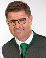 Ein lächelnder Mann mit Brille, der eine graue Weste über einem weißen Hemd und einer grünen Krawatte trägt, posiert für ein Bild.