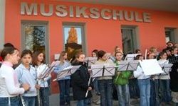 Eine Gruppe junger Menschen steht vor einem Gebäude mit der Aufschrift 'Musikschule' und hält Musikinstrumente in den Händen. Sie scheinen sich auf eine Aufführung vorzubereiten.