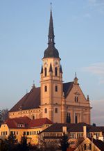 Eine große Kirche mit einem hohen Turm, goldenen Akzenten und einer Uhr, umgeben von Gebäuden unter einem klaren blauen Himmel.