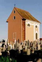 Eine kleine Kirche mit einem braunen Dach und beigefarbenen Wänden steht auf einem Friedhof, der mit vielen Grabsteinen und Kreuzen gefüllt ist, unter einem blauen Himmel.
