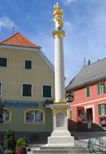 Eine hohe weiße Säule mit einer goldenen Statue auf der Spitze steht in der Mitte der Straße vor zwei Gebäuden mit gelben und roten Wänden.