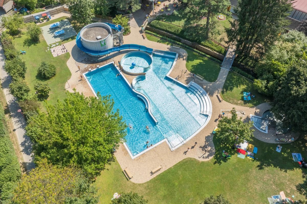 Eine Draufsicht auf einen großen Swimmingpool mit einer Wasserrutsche, umgeben von grünem Rasen und Bäumen. Mehrere Menschen schwimmen im Pool.