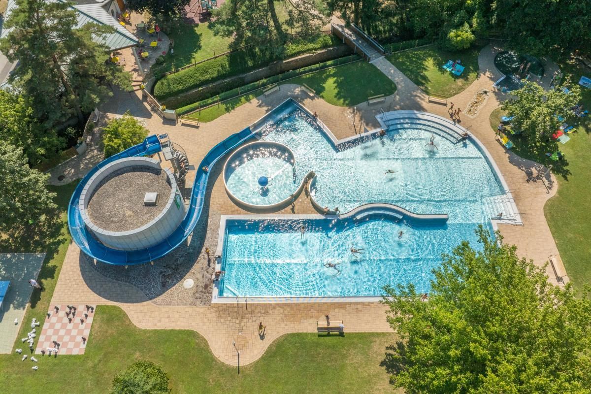 Ein Luftbild eines Resorts mit einem großen Schwimmbecken, das eine blaue Rutsche aufweist. Mehrere Menschen schwimmen im Becken, und es gibt Bäume und Gras in der Umgebung.