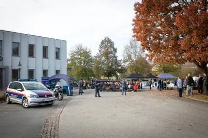 Eine Veranstaltung im Freien mit einem Polizeiauto, Zelten und Menschen, die in Stühlen vor einem Gebäude mit Herbstbäumen sitzen.