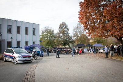Eine Veranstaltung im Freien mit einem Polizeiauto, Zelten und Menschen, die in Stühlen vor einem Gebäude mit Herbstbäumen sitzen.