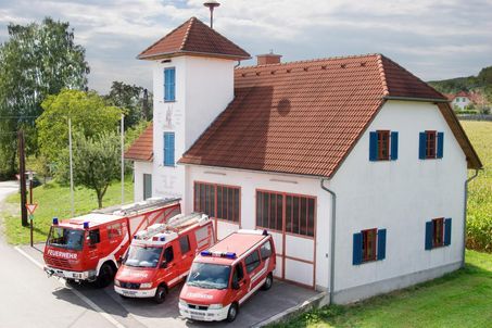 Ein Feuerwehrhaus mit rotem Dach und blauen Fensterläden ist zu sehen. Drei rote Rettungsfahrzeuge sind davor geparkt. Das Gebäude ist von Bäumen und Gras umgeben.