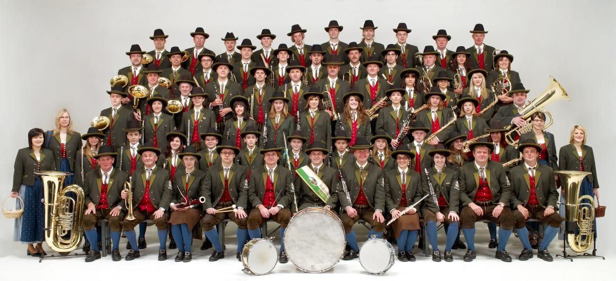 Eine Gruppe von Musikern in traditionellen grünen Uniformen und Hüten posiert für ein Foto mit ihren Instrumenten. Die Instrumente umfassen Trompeten, Posaunen, Flöten und Trommeln. Sie alle lächeln und schauen in die Kamera. Das Foto wurde in einem Studio mit weißem Hintergrund aufgenommen.