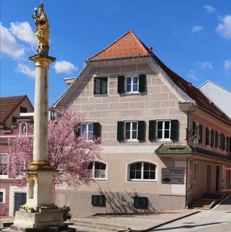 Eine goldene Statue auf einer Säule steht vor einem beigen Gebäude mit rotem Dach, geschmückt mit mehreren Fenstern und einem Schild. In der Nähe blüht ein rosa Baum.