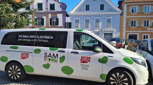 Ein weißer Van mit grünen Punkten steht vor einem Gebäude. Es steht 'Retter Reisen' und hat ein Logo. Der Van ist 100% elektrisch.