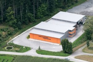 Eine Vogelperspektive auf ein großes Industriegebäude mit grauem Dach und orangefarbenen Wänden. Es ist von Bäumen umgeben und hat einen gepflasterten Bereich davor. Ein Auto ist am Straßenrand geparkt.