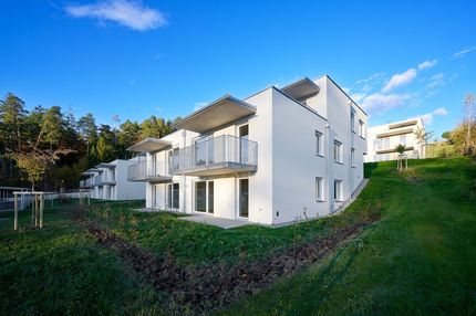 Ein modernes weißes Apartmenthaus mit Balkonen ist in einen Hang eingebettet, umgeben von Grün und Bäumen unter einem blauen Himmel.