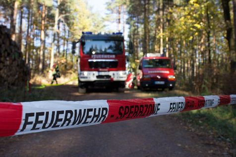Zwei rote Feuerwehrfahrzeuge sind auf einer unbefestigten Straße geparkt, mit einem Feuerwehrmann in der Nähe. Das Gebiet ist von Bäumen umgeben. Ein Absperrband mit den Worten 'Feuerwehr' und 'Sperrzone' ist über die Straße gelegt.