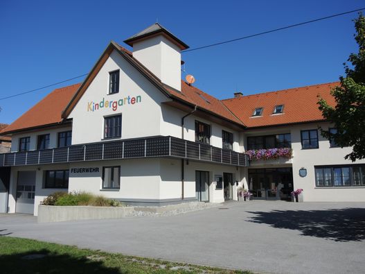 Ein weißes Gebäude mit rotem Dach und Ziegelwänden. Es steht 'Kindergarten' auf der Vorderseite. Es gibt einen Balkon mit Solarmodulen und ein Feuerwehrschild. Blumen stehen vor dem Eingang.