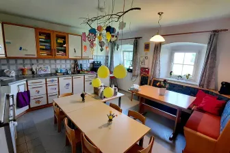 Eine helle, fröhliche Küche mit einem Essbereich. Tische und Stühle sind mit gelben Luftballons darüber angeordnet. Dekorationen und Blumen schmücken die Tische, und der Raum verfügt über Schränke, ein Waschbecken und ein Fenster.