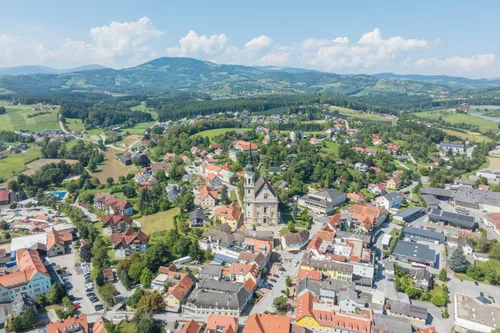 Luftaufnahme einer Stadt mit einer Kirche und vielen Gebäuden, umgeben von Bäumen und Bergen. Die Stadt liegt auf dem Land mit klarem blauem Himmel.