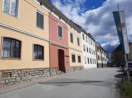 Eine leere Straße mit einem Gebäude mit der Aufschrift 'Marktgemeindeamt'. Das Gebäude hat eine rote und rosa Wand und eine Steinmauer als Basis.