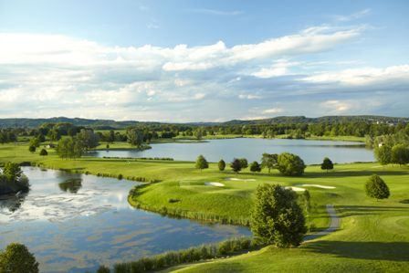 Luftaufnahme eines Golfplatzes mit einem Fluss, Bäumen und einem See. Der Himmel ist teilweise bewölkt und die Sonnenstrahlen beleuchten die Landschaft.