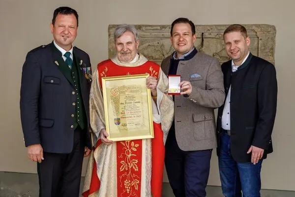 Ein Mann in roten Gewändern hält ein gerahmtes Dokument in der Hand, flankiert von drei anderen Männern. Zwei sind in Geschäftskleidung, einer mit einem Lächeln, und einer in Uniform. Sie stehen vor einer Steinmauer.