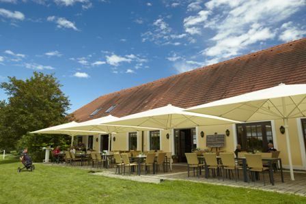 Außenbereich mit Tischen und Stühlen unter großen Sonnenschirmen in einem Golfclub an einem sonnigen Tag.