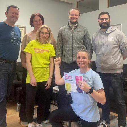 Eine Gruppe von sechs Personen steht lächelnd zusammen in einem Raum. Die Frau vorne hält ein Flugblatt. Die anderen tragen Freizeitkleidung. Der Hintergrund besteht aus einer Wand und einer Couch.