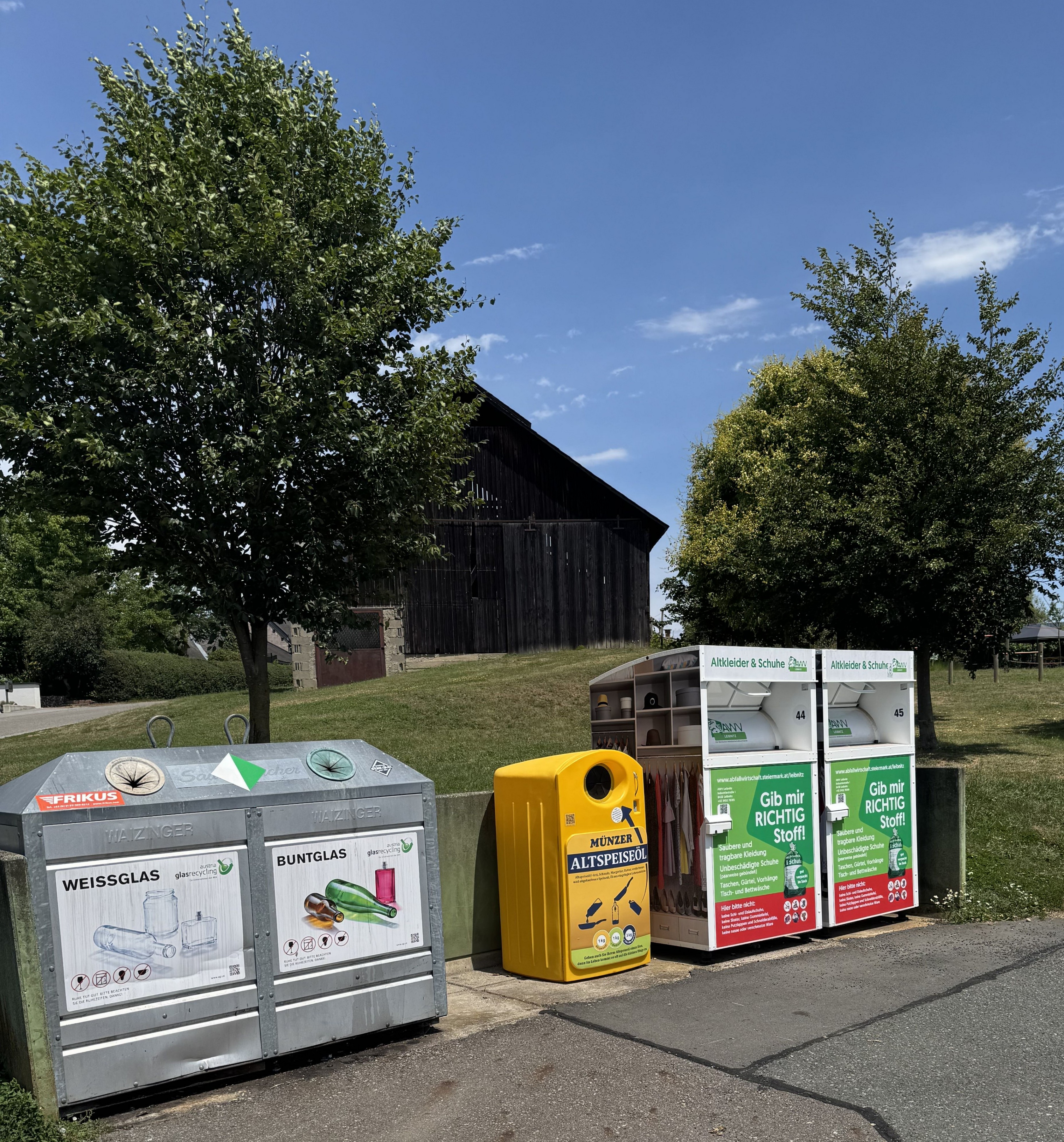 Eine Reihe von Recyclingbehältern auf einem Grasfeld mit einem Scheunen im Hintergrund. Es gibt zwei Behälter für Glas, einen für gemischten Recycling und einen für gefährlichen Abfall.