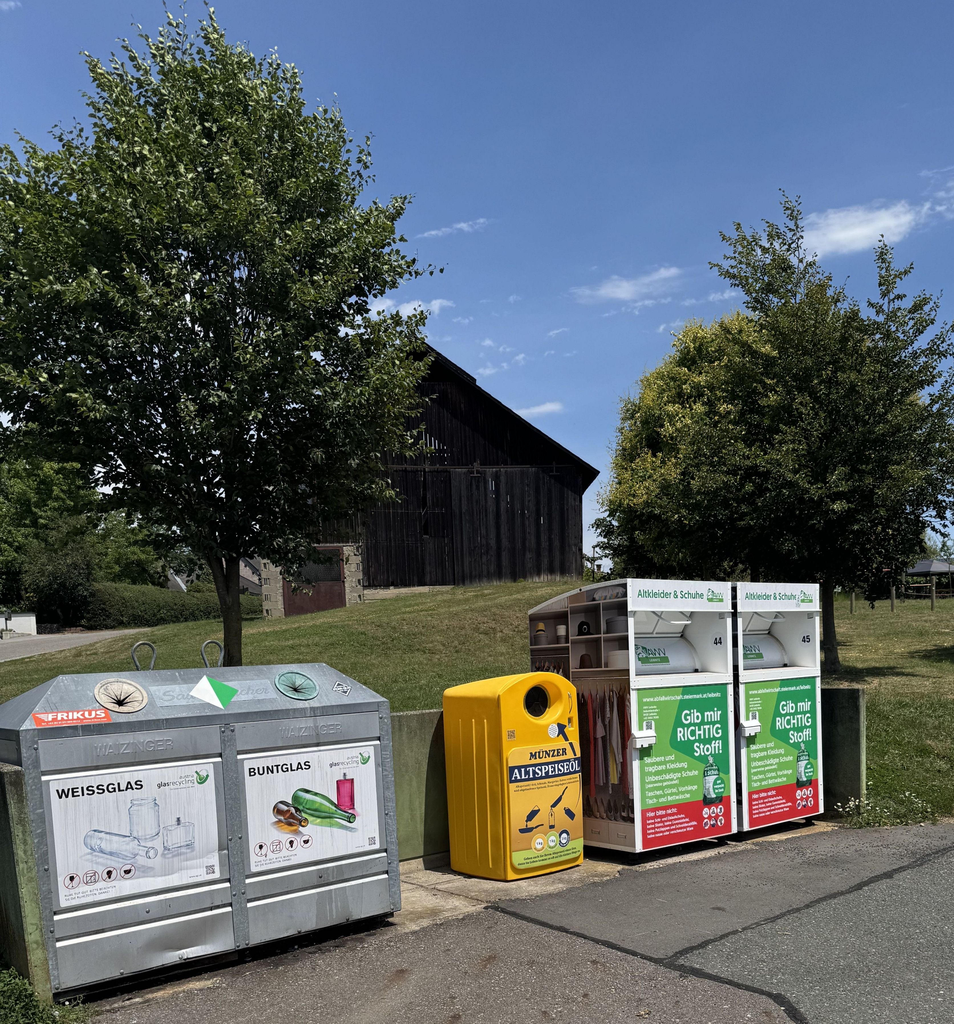 Eine Reihe von Recyclingbehältern auf einem Grasfeld mit einem Scheunen im Hintergrund. Es gibt zwei Behälter für Glas, einen für gemischten Recycling und einen für gefährlichen Abfall.