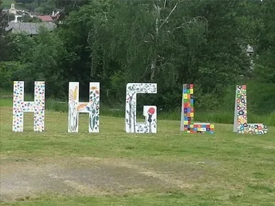 Weiße Buchstaben, die HGHL bilden, sind auf einer grünen Wiese platziert. Die Buchstaben bestehen aus farbenfrohen Mosaikfliesen. Im Hintergrund sind Bäume und Häuser zu sehen.