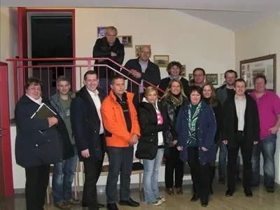 Eine Gruppe von Menschen, sowohl Männer als auch Frauen, posiert für ein Foto in einem Gebäude. Sie tragen Winterkleidung und stehen auf einem Holzboden.