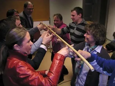 Eine Gruppe von Menschen in einem Raum halten sich an einem langen Stock die Hände. Einige tragen formelle Kleidung, andere lässige Kleidung. Die Frau in der roten Lederjacke lächelt.