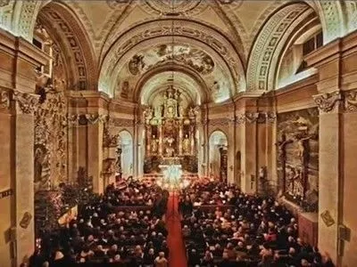 Eine große Gemeinde ist in einer großen Kathedrale versammelt, mit aufwendigen Verzierungen, prächtigen Säulen und einem zentralen Altar, geschmückt mit Kerzen. Die Atmosphäre ist feierlich und ehrfürchtig.