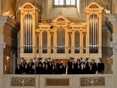 Ein Chor steht vor einer aufwendig verzierten Orgel in einem großen, klassischen Saal, wobei natürliches Licht von den Fenstern oben hereinfällt.