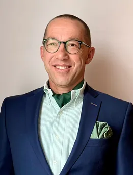 Ein Mann mit Brille, einem blauen Anzug und einem grünen Hemd mit einem grünen Einstecktuch lächelt.