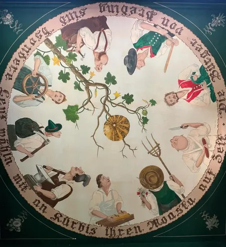 Ein kreisförmiges Wandbild zeigt verschiedene Männer in traditioneller Kleidung bei unterschiedlichen Tätigkeiten. Eine goldene Gurke befindet sich im Zentrum, umgeben von Figuren, die Werkzeuge wie einen Hammer und eine Sichel halten. Der Rand hat eine Inschrift.