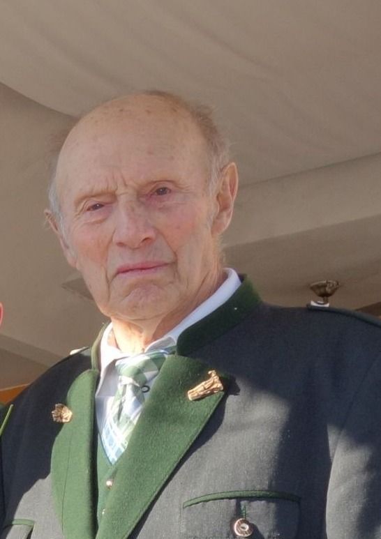 Ein älterer Mann mit ernster Miene trägt eine grün-graue Jacke mit einem weißen Hemd darunter.