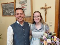 Ein Mann und eine Frau stehen lächelnd nebeneinander, der Mann in einer Weste und die Frau mit einem Blumenstrauß. Sie stehen vor einer Wand mit einem gerahmten Dokument und einem Kruzifix.