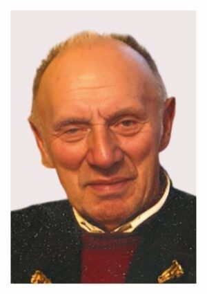 Ein älterer Mann mit ernster Miene, der eine schwarze Jacke und einen roten Pullover trägt. Er hat kurzes, graues Haar und schaut direkt in die Kamera.
