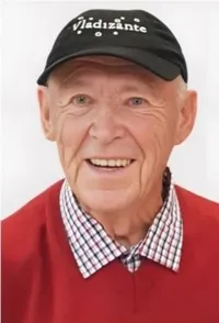 Ein älterer Mann mit einer schwarzen Kappe mit weißem Text und einem roten Pullover mit kariertem Kragen, lächelt in die Kamera.