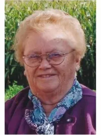 Porträt einer älteren Frau mit kurzen blonden Haaren, die eine lila Jacke und einen blauen Schal trägt. Sie trägt eine Brille und lächelt vor einem verschwommenen Hintergrund aus grünen Pflanzen.
