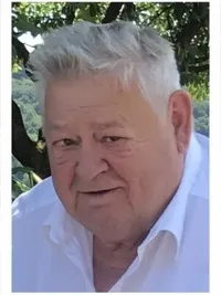 Ein älterer Mann mit weißen Haaren und einem weißen Hemd steht draußen unter einem Baum. Er schaut nach rechts und lächelt leicht.