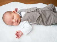 Ein Babyjunge in einer grauen Weste und Fliege schläft friedlich auf einer weißen Decke, seine Hände neben sich.