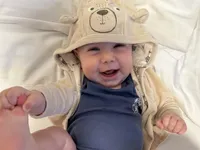 Ein Baby in einer Bären-Kapuzenjacke liegt auf einem weißen Bett und lächelt, eine Hand erhoben.