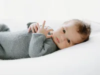 Ein Baby in einem grauen gestrickten Pullover liegt auf einer weißen Oberfläche und blickt neugierig mit einem leichten Lächeln zur Kamera.