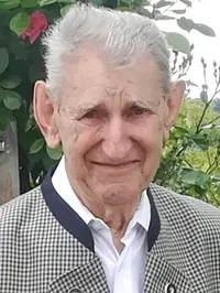 Ein älterer Mann mit weißem Haar und einem Lächeln, der eine karierte Jacke und ein weißes Hemd trägt, steht in einem Garten mit grünen Pflanzen im Hintergrund.