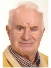 Ein älterer Mann mit weißem Haar und fröhlicher Miene, der einen gelben Pullover und ein kariertes Hemd trägt.