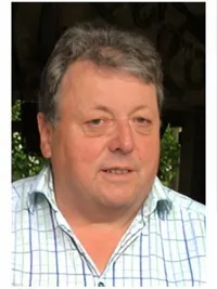 Ein Mann mit grauem Haar, der ein hellblau kariertes Hemd trägt, posiert für ein Foto vor grünem Hintergrund.