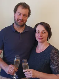 Ein Mann und eine Frau stehen eng beieinander, lächeln und halten Sektgläser mit Champagner. Der Mann trägt ein blaues Poloshirt, und die Frau hat kurze Haare und ein gemustertes Oberteil. Sie scheinen für ein Foto vor einer schlichten weißen Wand zu posieren.
