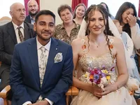 Eine Hochzeitszeremonie mit dem Bräutigam und der Braut, umgeben von der Familie. Die Braut trägt ein mit Blumen geschmücktes Kleid und hält einen Blumenstrauß. Der Bräutigam trägt einen blauen Anzug und eine Blumenkrawatte. Hinter ihnen stehen andere, einschließlich einer Frau mit einem Baby.